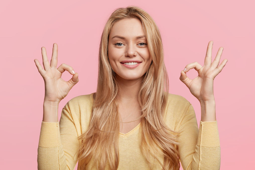 Glad attractive woman shows ok sign as expresses approval, gestures in studio against pink background, has cheerful expression. Photo of beautiful female has appealing appearance, being optimstic