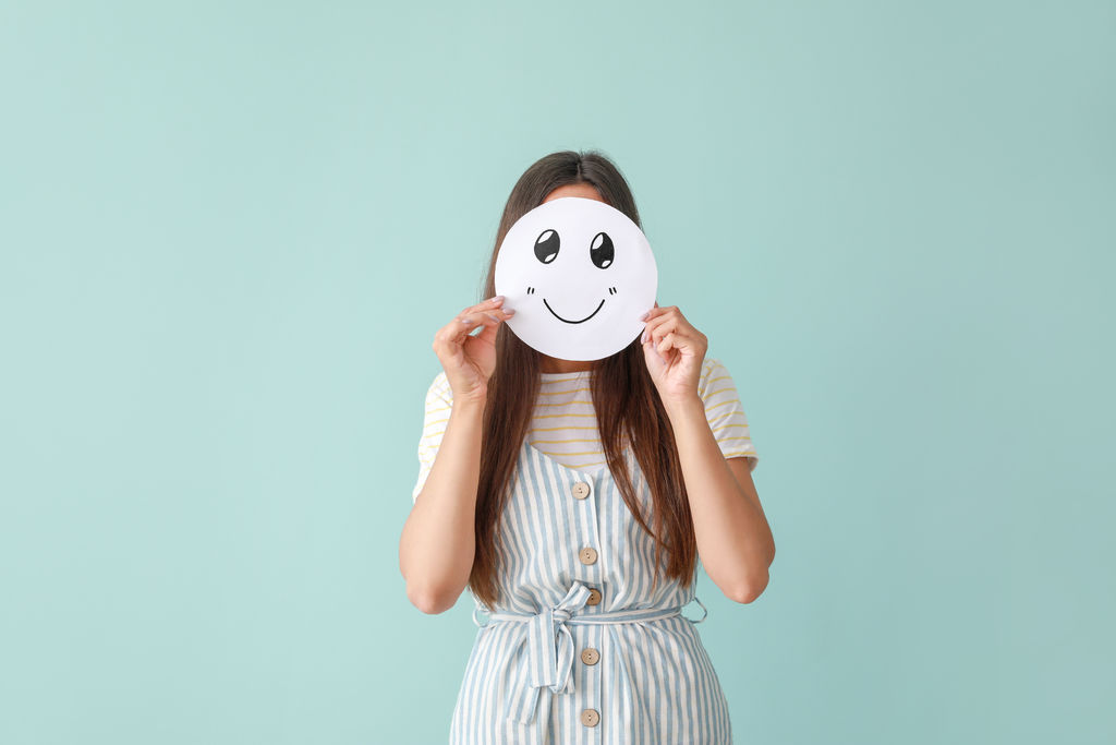 Woman hiding face behind sheet of paper with drawn emoticon on color background