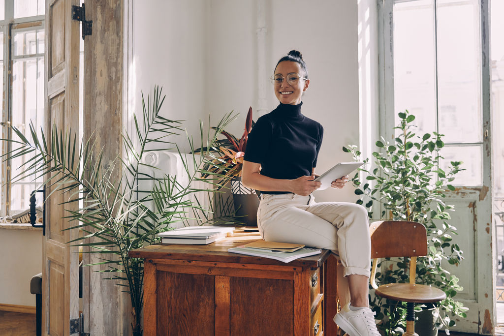 Happy young woman holding digital tablet while sitting on the desk in office