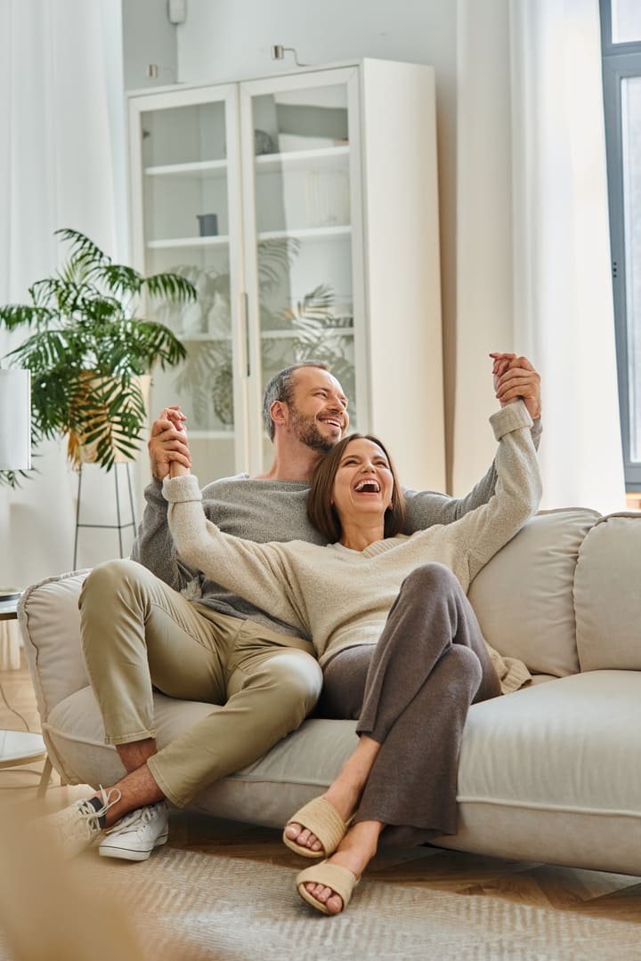 excited child-free couple holding hands and laughing on cozy couch in living room, fun and leisure