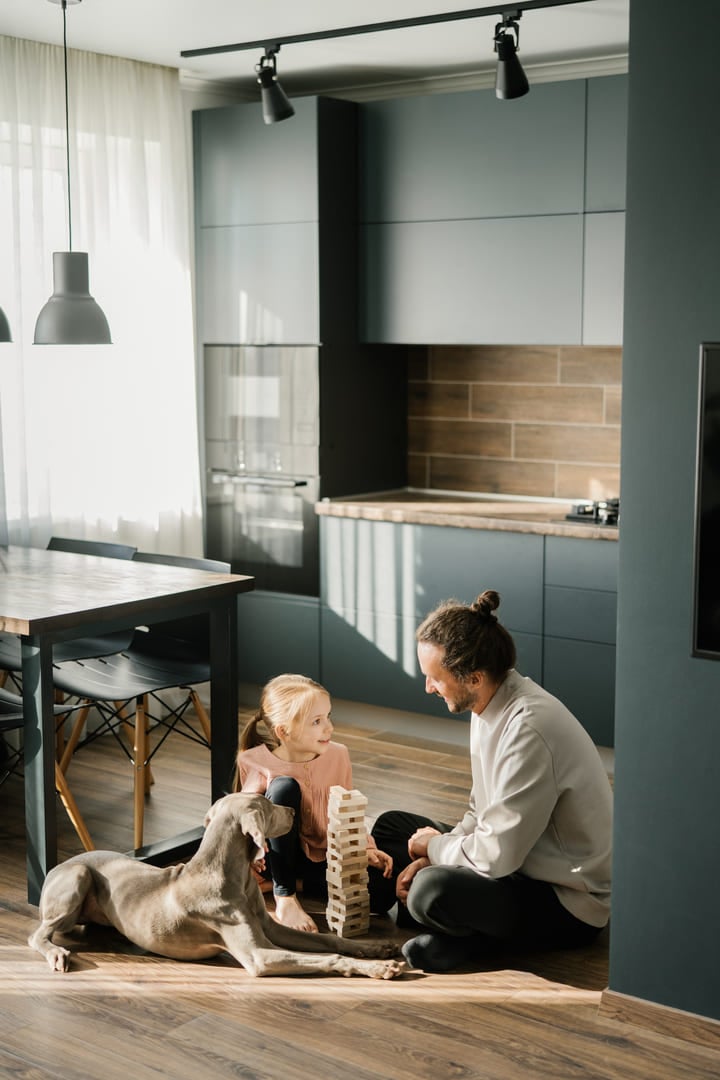 Dad and daughter are playing a board game in the kitchen, sitting on the floor. A dog is lying nearby. Scandinavian and high-tech style in the interior.