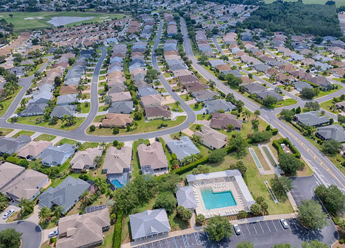 Aerial view of a master-planned residential community with curved streets, single-story homes, landscaped lawns, and a community pool, surrounded by greenery
