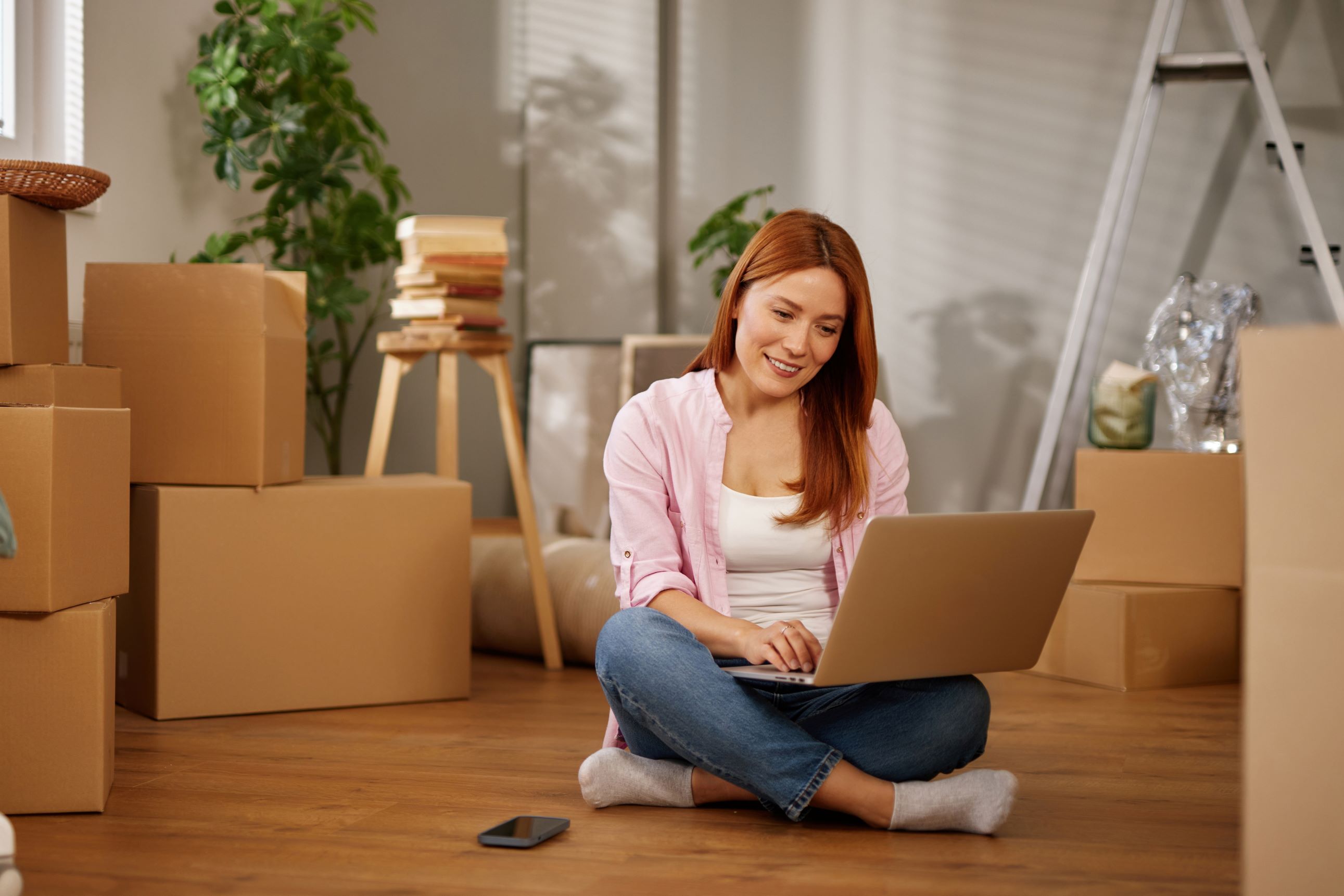 A woman sits cross-legged on the floor of her new living space, focused on her laptop while surrounded by unpacked boxes, enjoying a moment of transition and planning her new life.