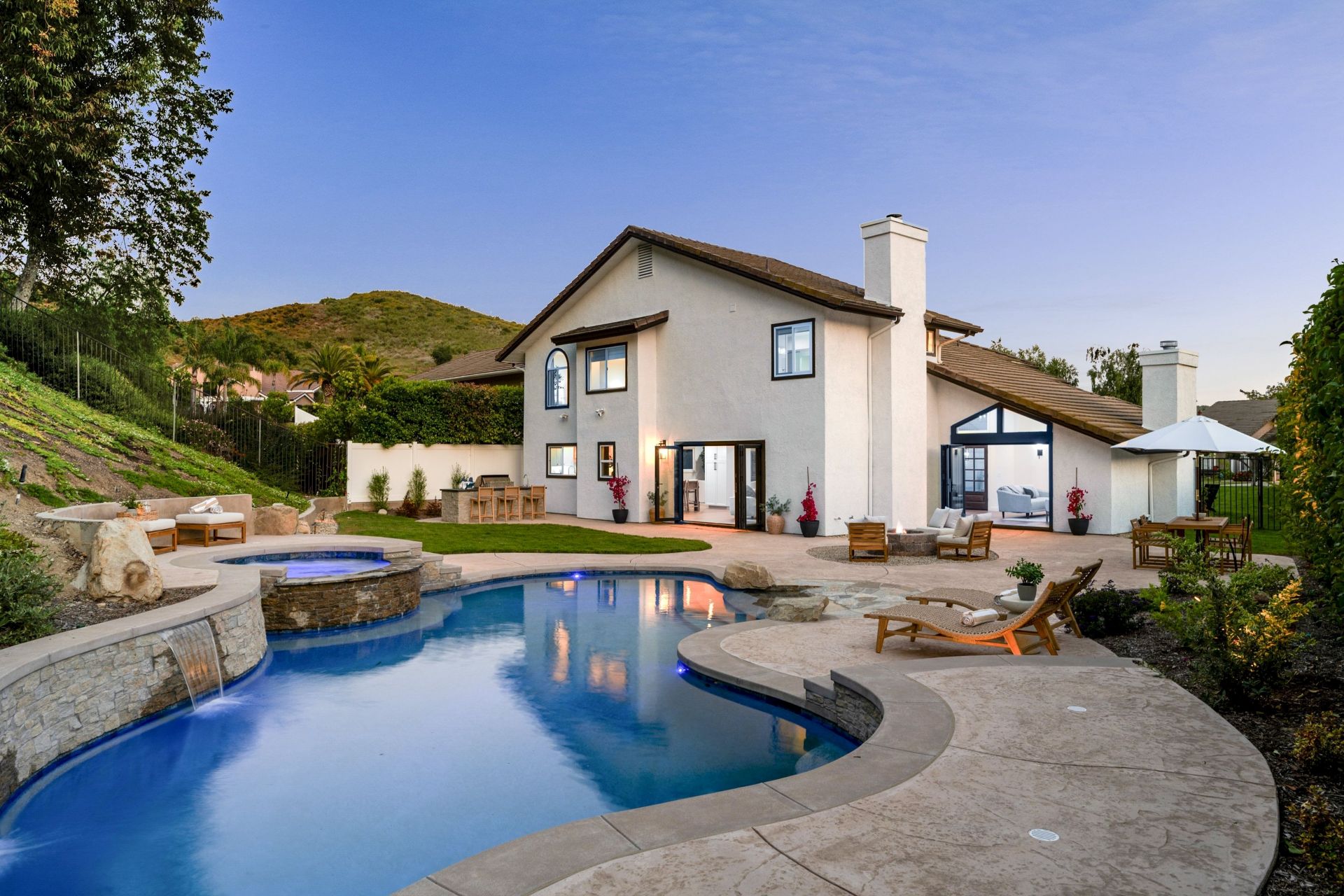 Modern luxury house with a swimming pool and patio area, surrounded by greenery and a clear evening sky.