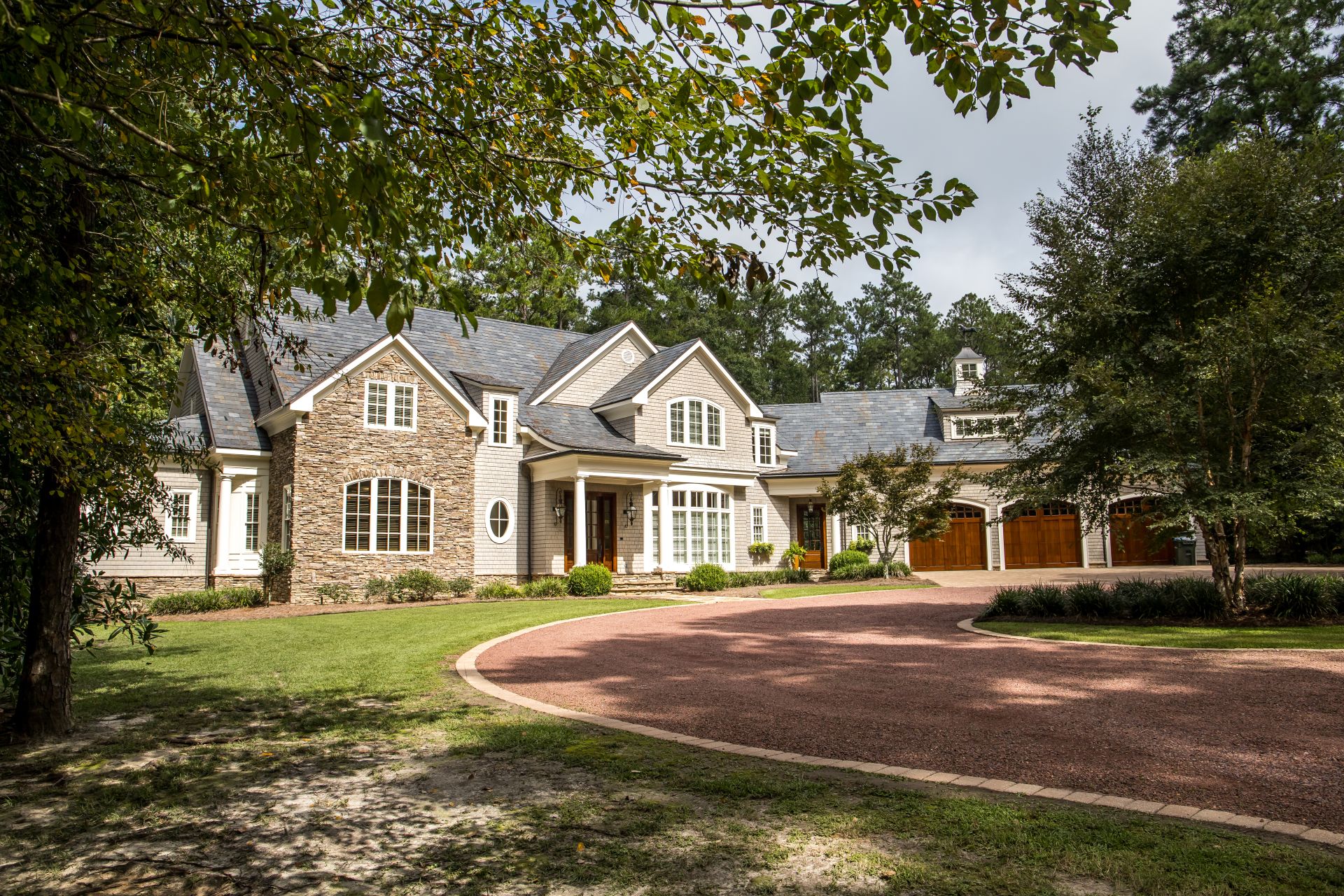 Front view of large estate home in the south with a gravel driveway and lots of windows. house made of brick, stone and clapboard in a cape cod style. and a triple garage with curb appeal