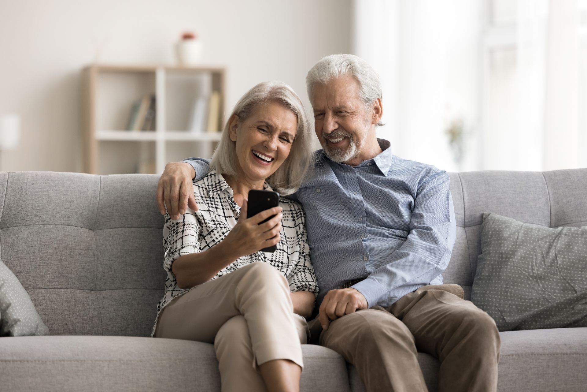 Happy aged wife and husband sit on sofa look at mobile phone screen, laughing, watching funny videos