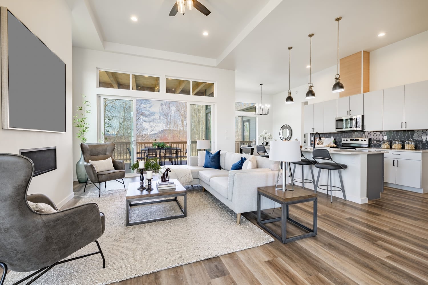 open concept living room with stylish furniture electric fireplace grey leather arm chairs windows revealing blue sky hardwood floors and view to kitchen (1)