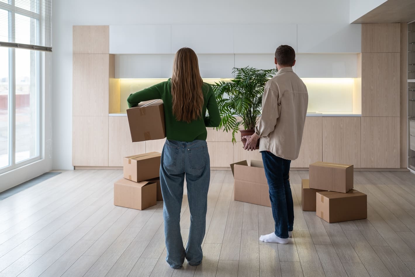 New home owners with boxes and houseplant in hands, rear view. Family couple taking things to another rented light apartment. Housewarming in mortgage apartment. Beginning of happy married life.