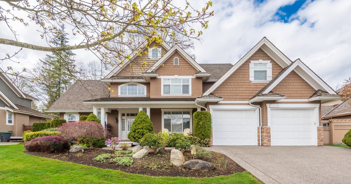 Two-story suburban house with a landscaped front yard, mature trees, and an attached double garage on a sunny day