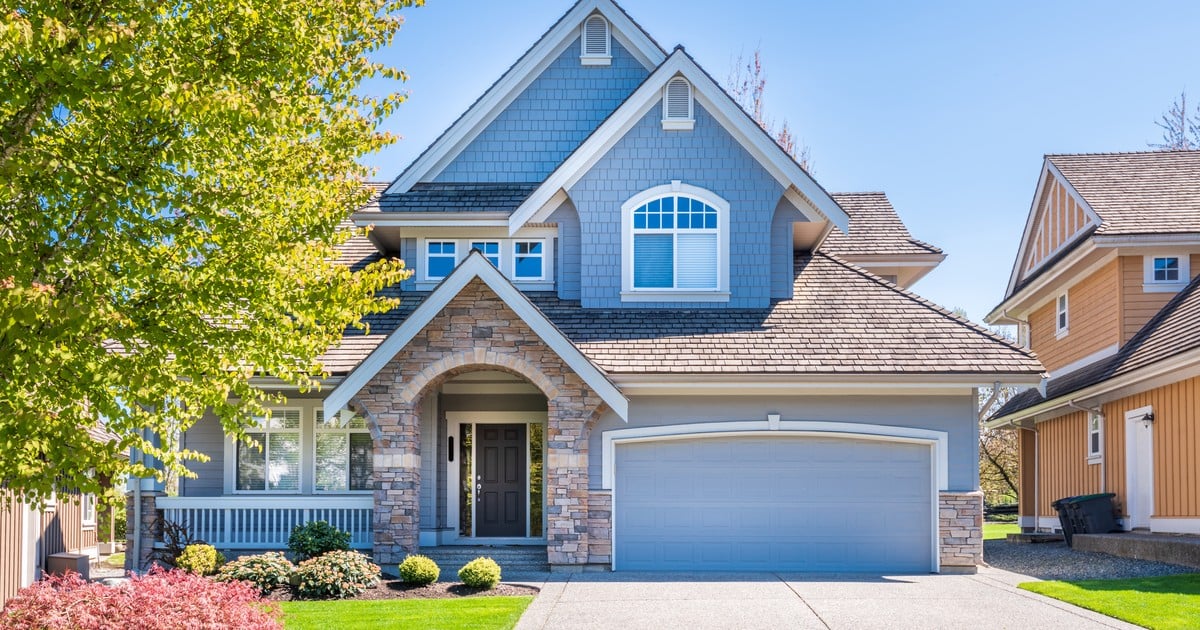 Two-story blue suburban house with stone archway entrance, an attached double garage, mature landscaping, and a paved driveway on a bright sunny day.