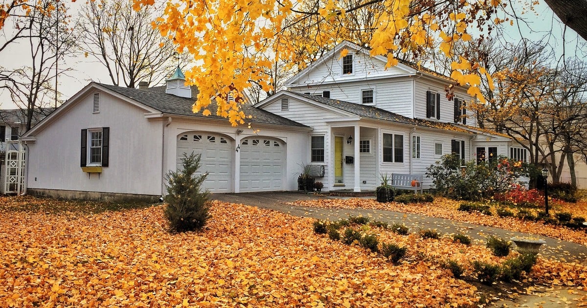 White two-story house with an attached garage, framed by a tree dropping golden autumn leaves, with a front lawn and walkway completely covered in fallen leaves.