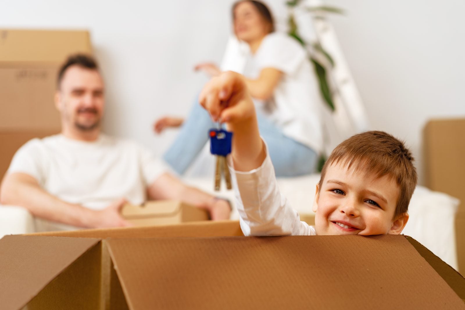Young boy smiling and holding up house keys from a moving box while his parents relax among boxes in their new home