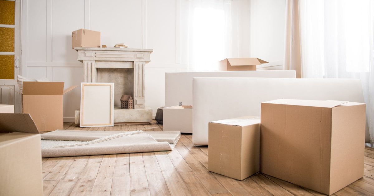 Cardboard moving boxes, a rolled-up rug and a picture frame in a bright, empty living room with a fireplace and wood floors.
