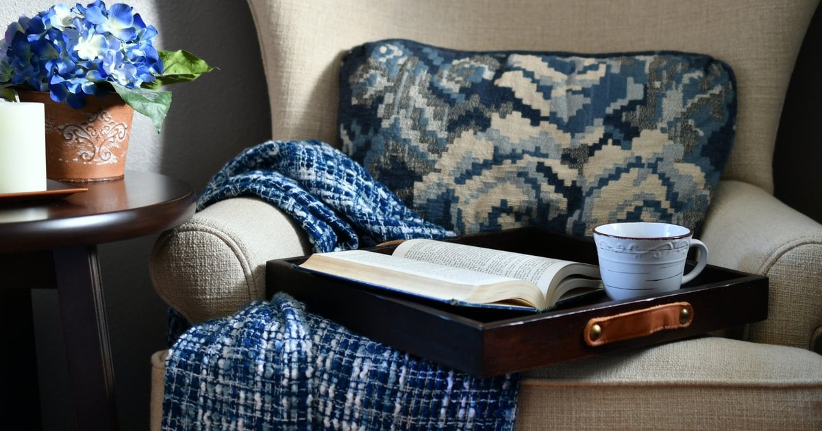 A cozy armchair with a blue patterned cushion, a knit blanket, a tray with an open book and a coffee mug, and blue hydrangeas on a side table.