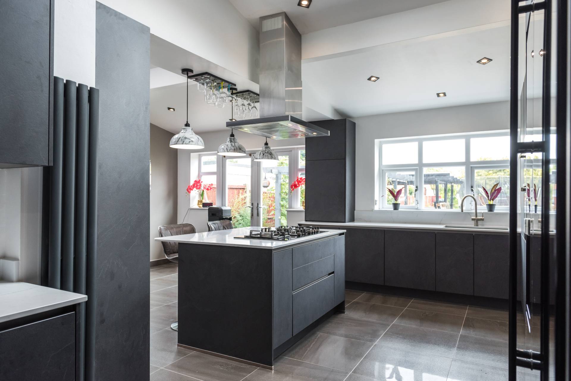 Modern minimalist kitchen with dark gray cabinets, a central island cooktop and stainless steel range hood, large windows letting in natural light, and sleek tile flooring.