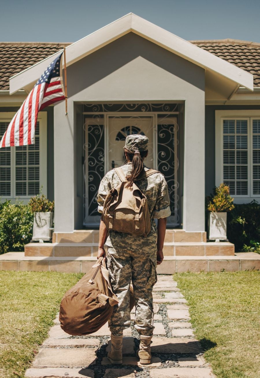 Rearview of a patriotic female soldier standing in front of her home with her luggage. Courageous servicewoman coming back home after serving her country in the military vertical