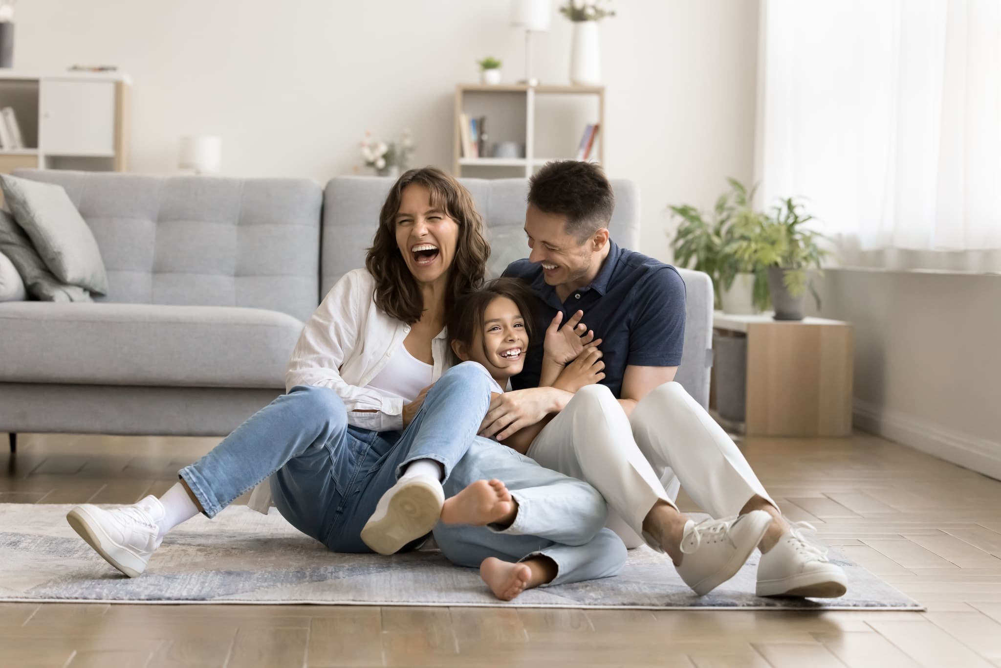 Cheerful excited parents tickling beloved kid girl, playing with child on heating floor in modern home interior, sitting on carpet, laughing, shouting for joy, enjoying family leisure