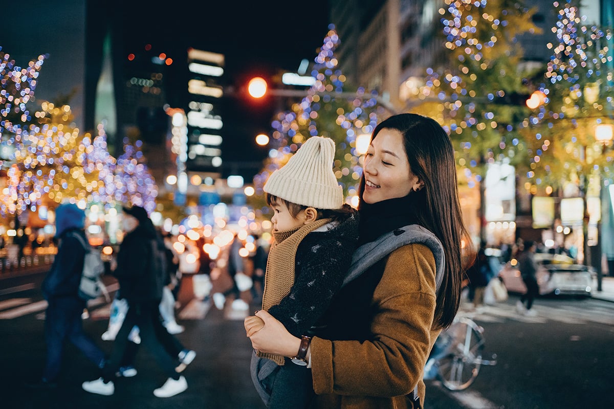 Young mother with little daughter walking in downtown city street at night, against illuminated and multi-coloured street lights and urban buildings