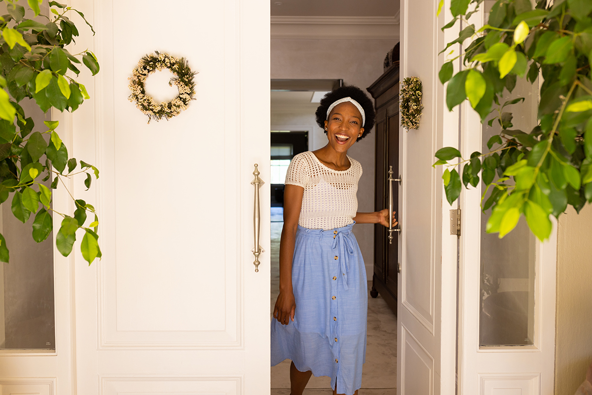 Cheerful african american mid adult woman with short afro hair opening door and standing at entrance. Unaltered, lifestyle, happy and welcoming concept.