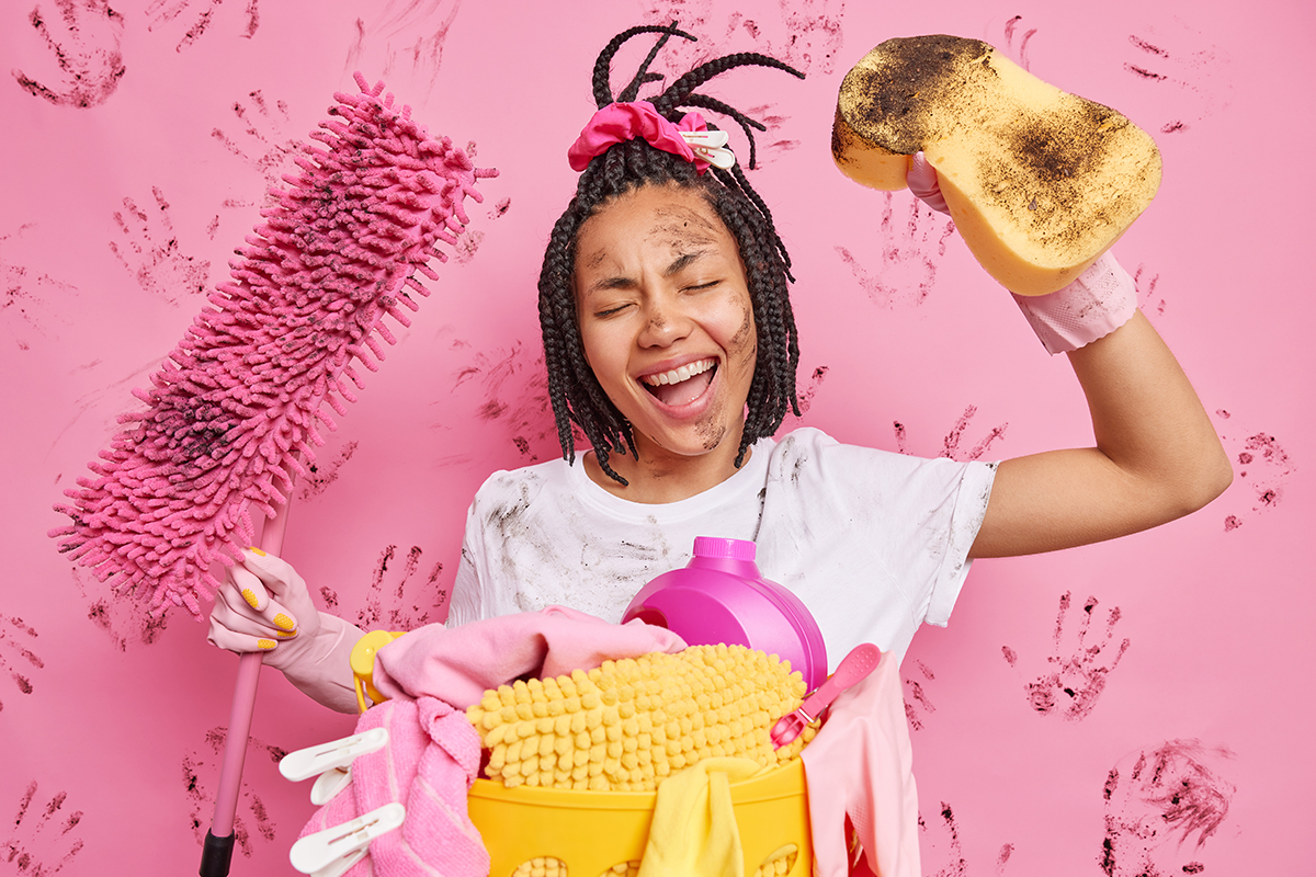 Joyful housewife with braided hair holds sponge and mop busy doing cleaning stands near basket of laundry and cleaning detergents wears dirty white t shirt rubber gloves isolated over pink background