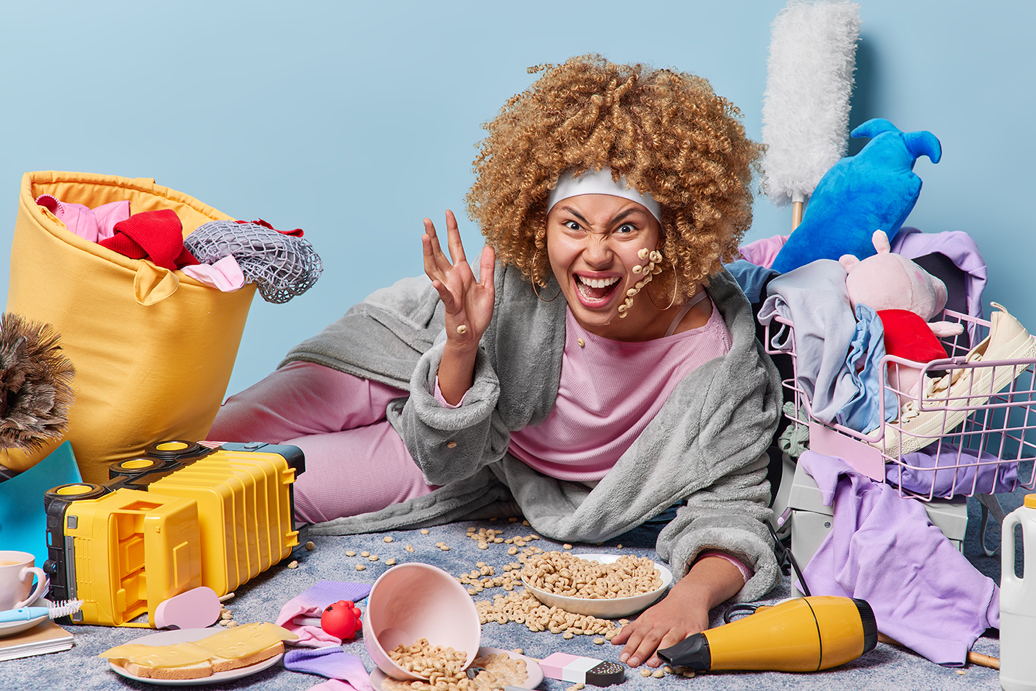 Crazy emotional woman with curly hair gestures and screams angrily wears domestic robe and headband surrounded by different items poses in messy room has to bring house in order. Lazy housewife