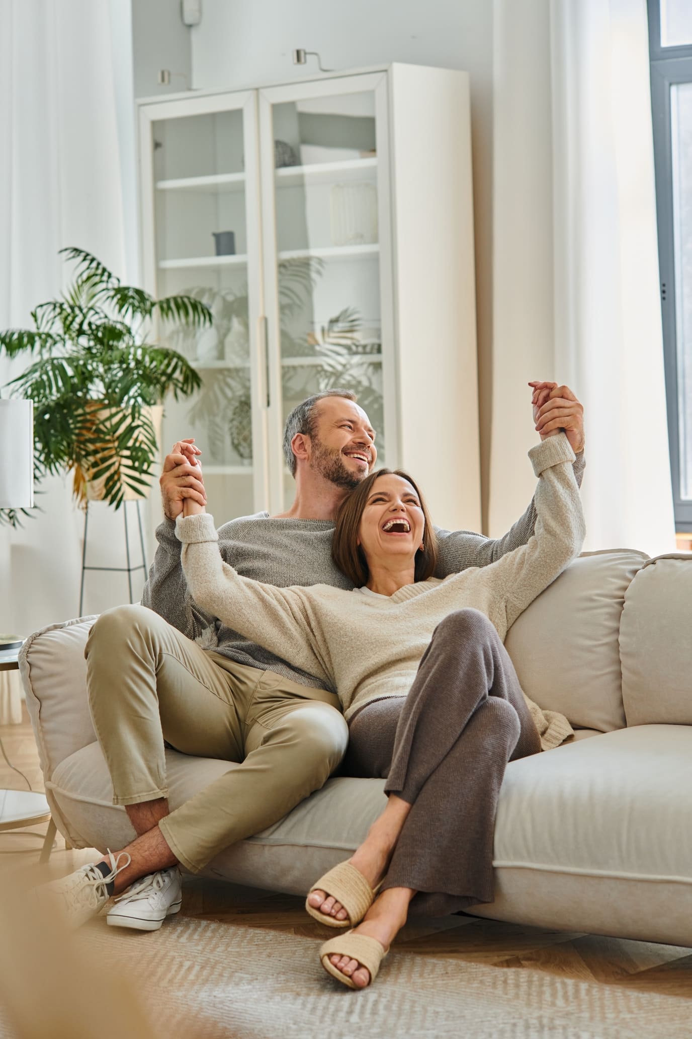 excited child-free couple holding hands and laughing on cozy couch in living room, fun and leisure