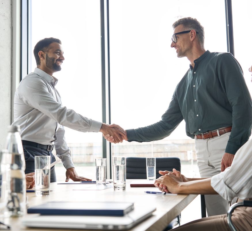 Two happy diverse professional business men executive leaders shaking hands after successful financial deal at group board office meeting