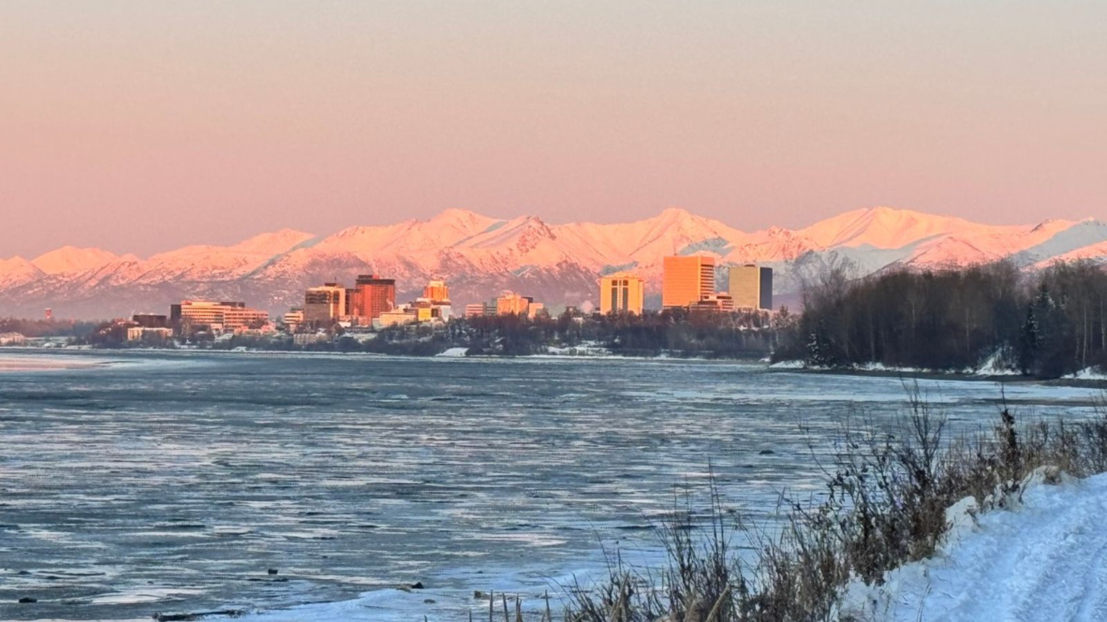 Downtown Anchorage skyline during winter sunset with snow-covered Chugach Mountains and frozen coastal water