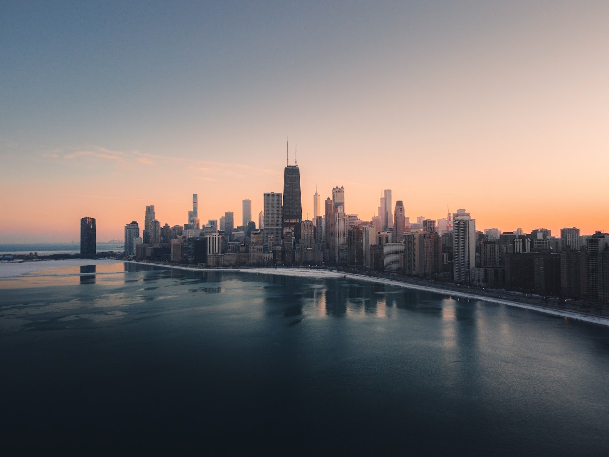 A panoramic aerial view of Chicago's cityscape in winter during sunset in Illinois, United States. It features buildings, snow, a lake, roads, a clear sky, and lake reflection.