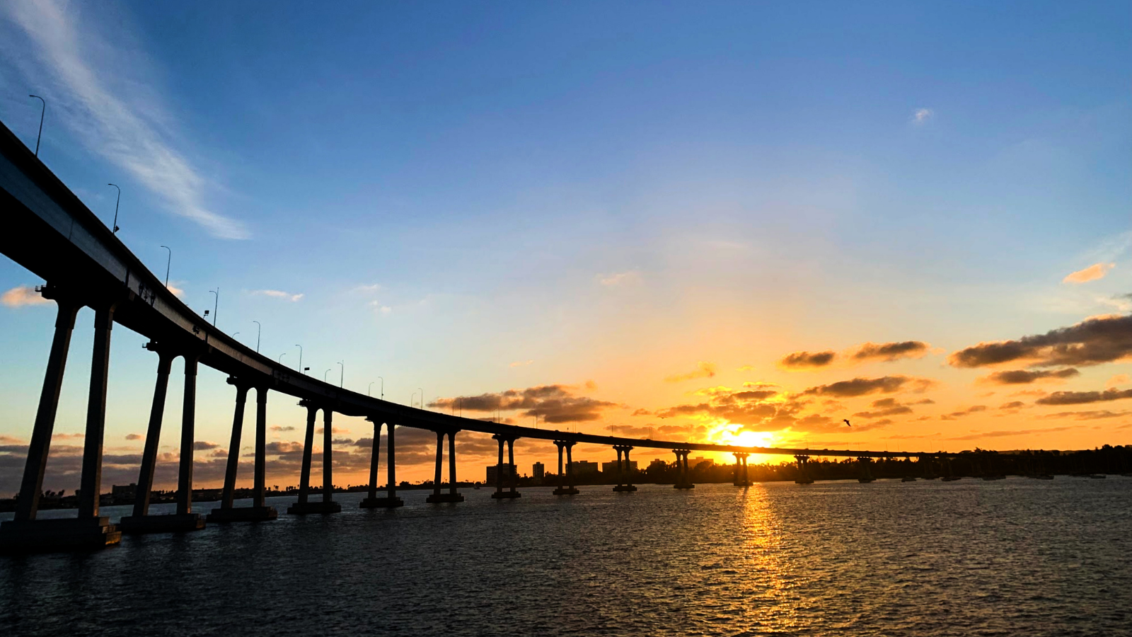 The Coronado Bridge silhouetted against an orange sunset over San Diego Bay, with the shoreline and city lights in the distance.