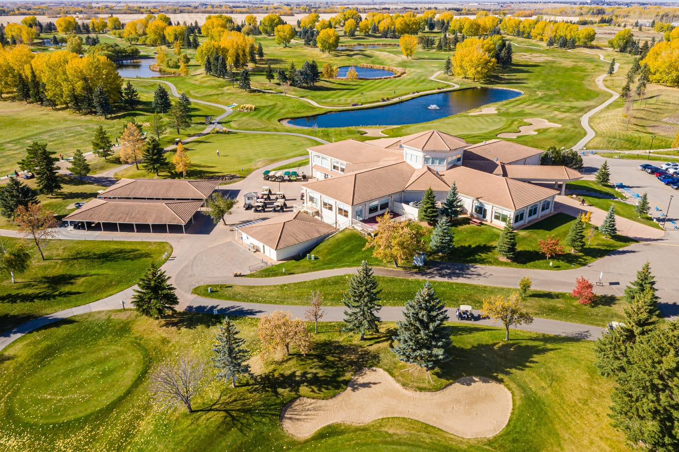 Golf course with a large building in the background. The building is white and has a red roof. The golf course is surrounded by trees and has a pond in the middle (1) (1)