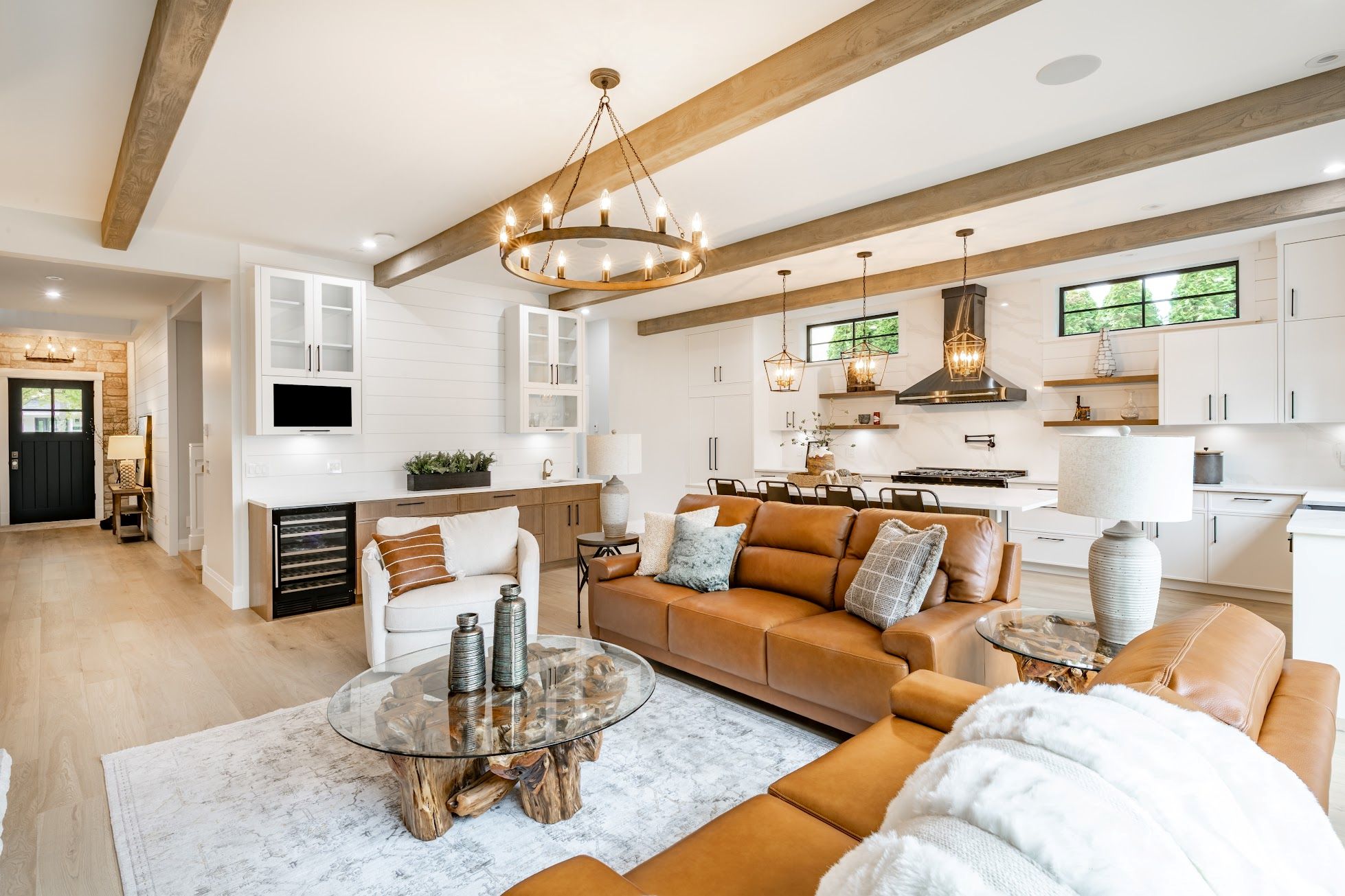 Living room interior with exposed wooden beams on the ceiling carmel colored leather sofa round wagon wheel chandelier fluffy white throw blanket hardwood floor view to kitchen and stone fire