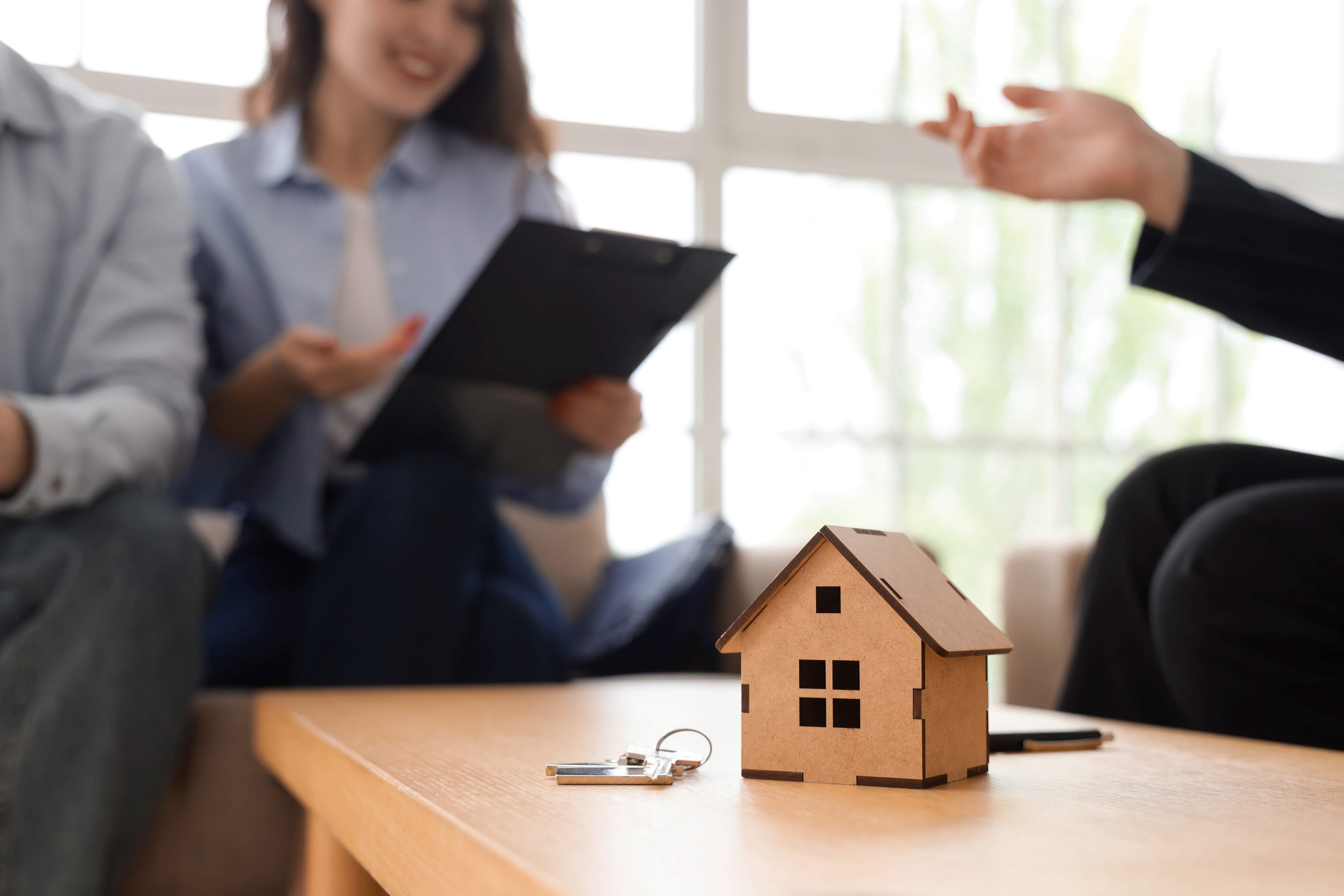 House figure and key on table against couple signing agreement with real estate agent in room, closeup