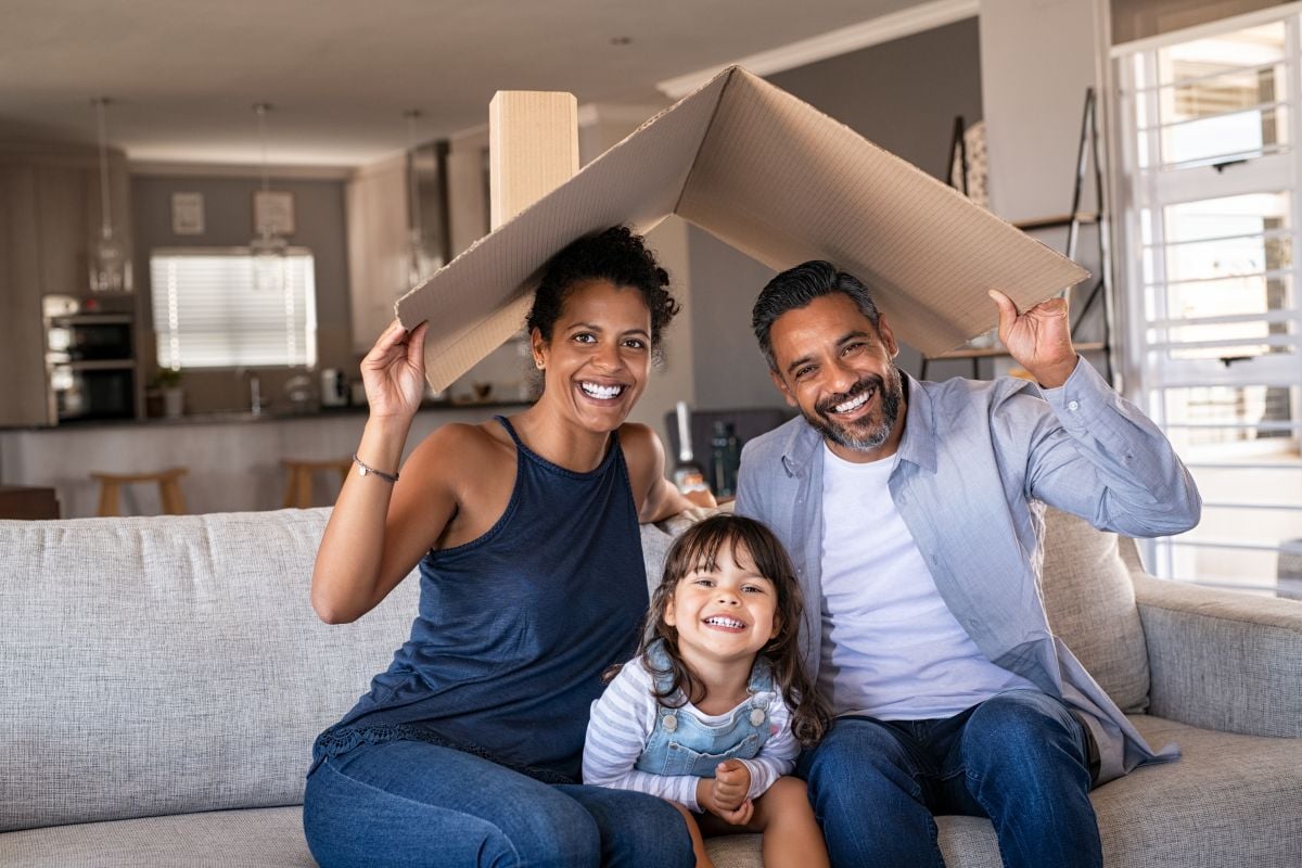 Portrait of smiling family sitting on couch holding cardboard roof and looking at camera.