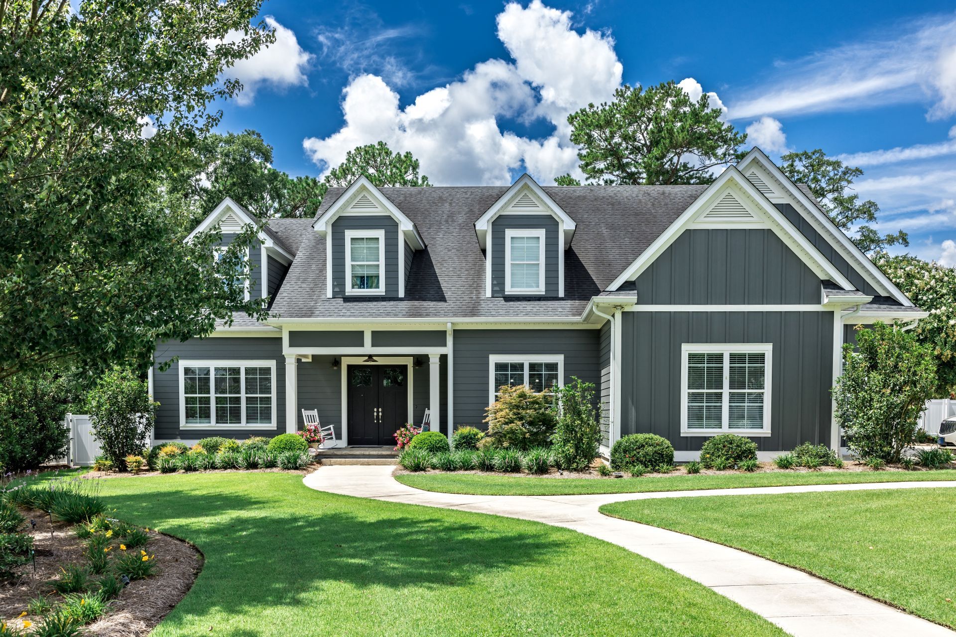 A large gray craftsman new construction house with a landscaped yard and leading pathway sidewalk on a sunny day with blue skies and clouds.