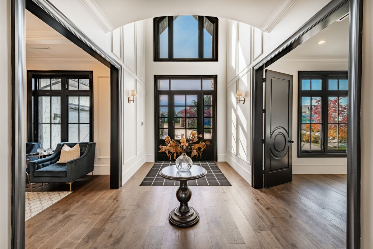 stately luxurious elegant entry hall foyer welcome front door with glass and large area rug round table with floral flower arrangement white wood panelled walls staircase stairs open concept
