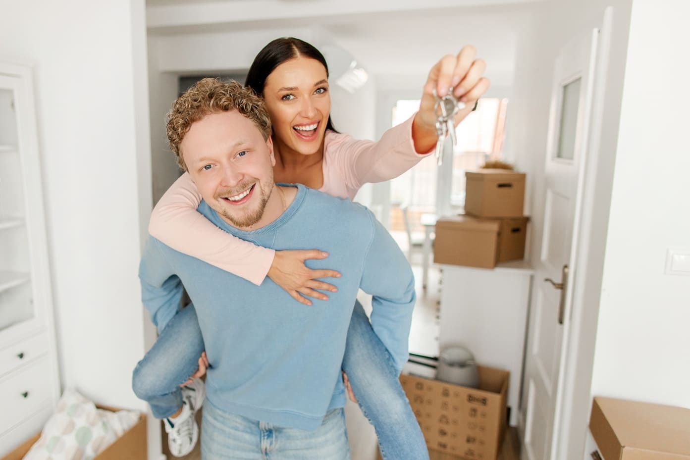 Excited lady getting piggyback ride from her husband and showing house key to camera, posing in new apartment on moving day (1) (1)