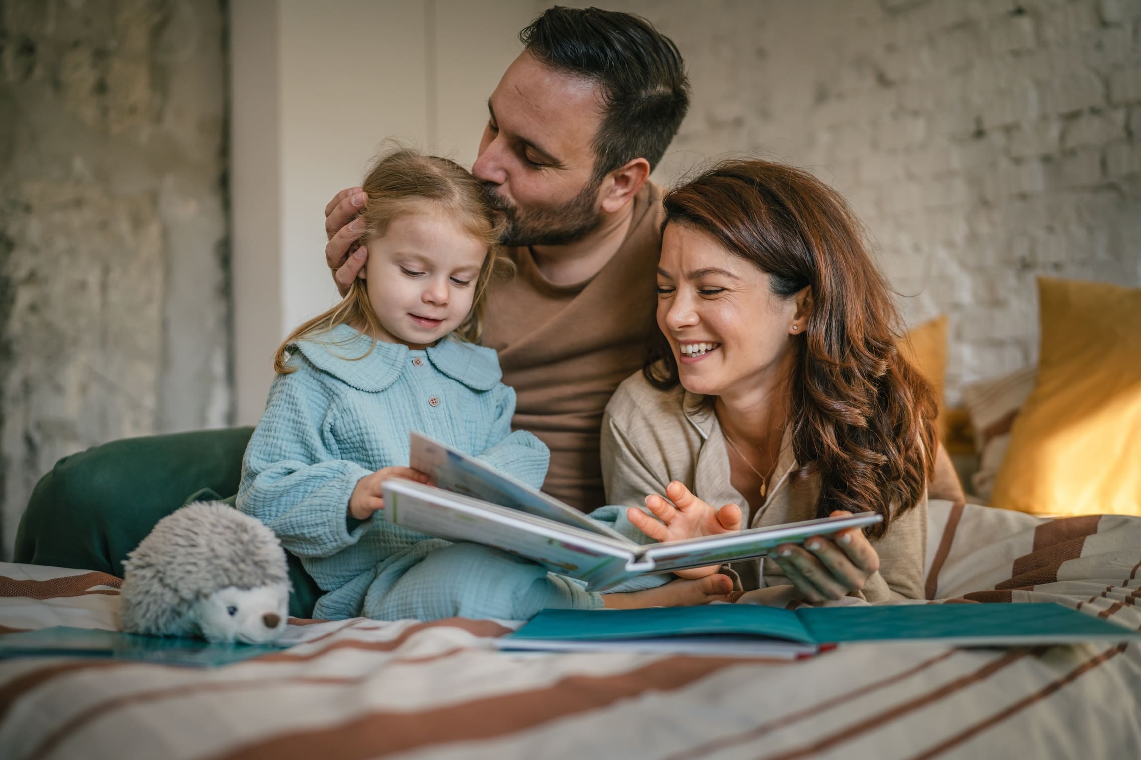 Happy parents and their young daughter are lying on a bed, bonding while reading a picture book and laughing aloud, sharing a joyful and intimate moment at home (1) (1)