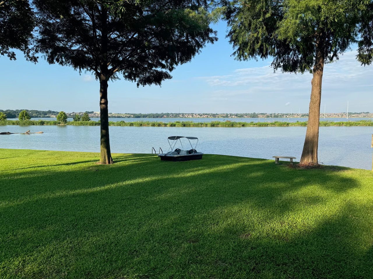 Grassy shoreline park on Lake Ray Hubbard with a pedal boat and bench under mature trees in Rowlett, TX
