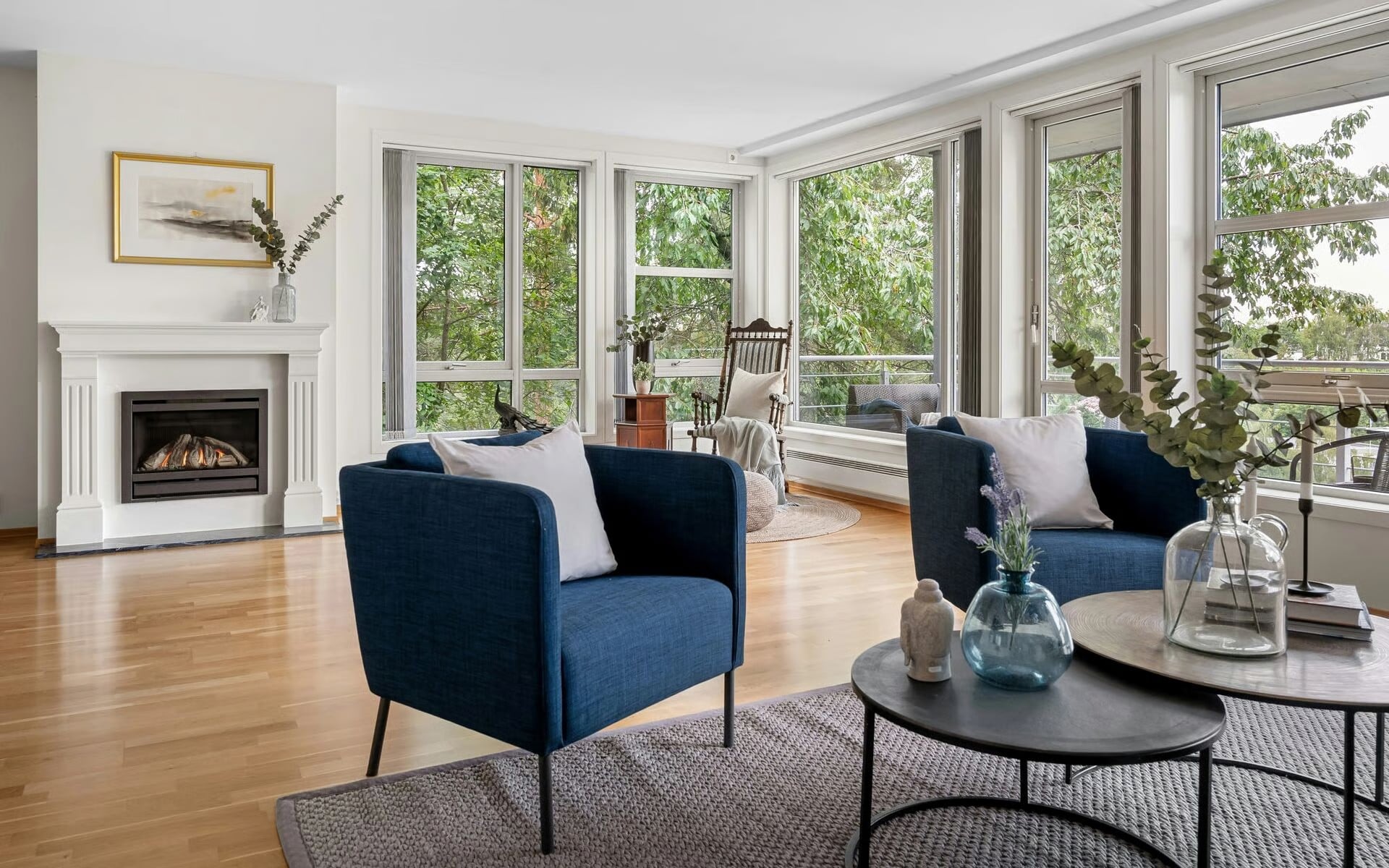 Living room with a white fireplace mantel, navy blue accent chairs, and large windows overlooking trees