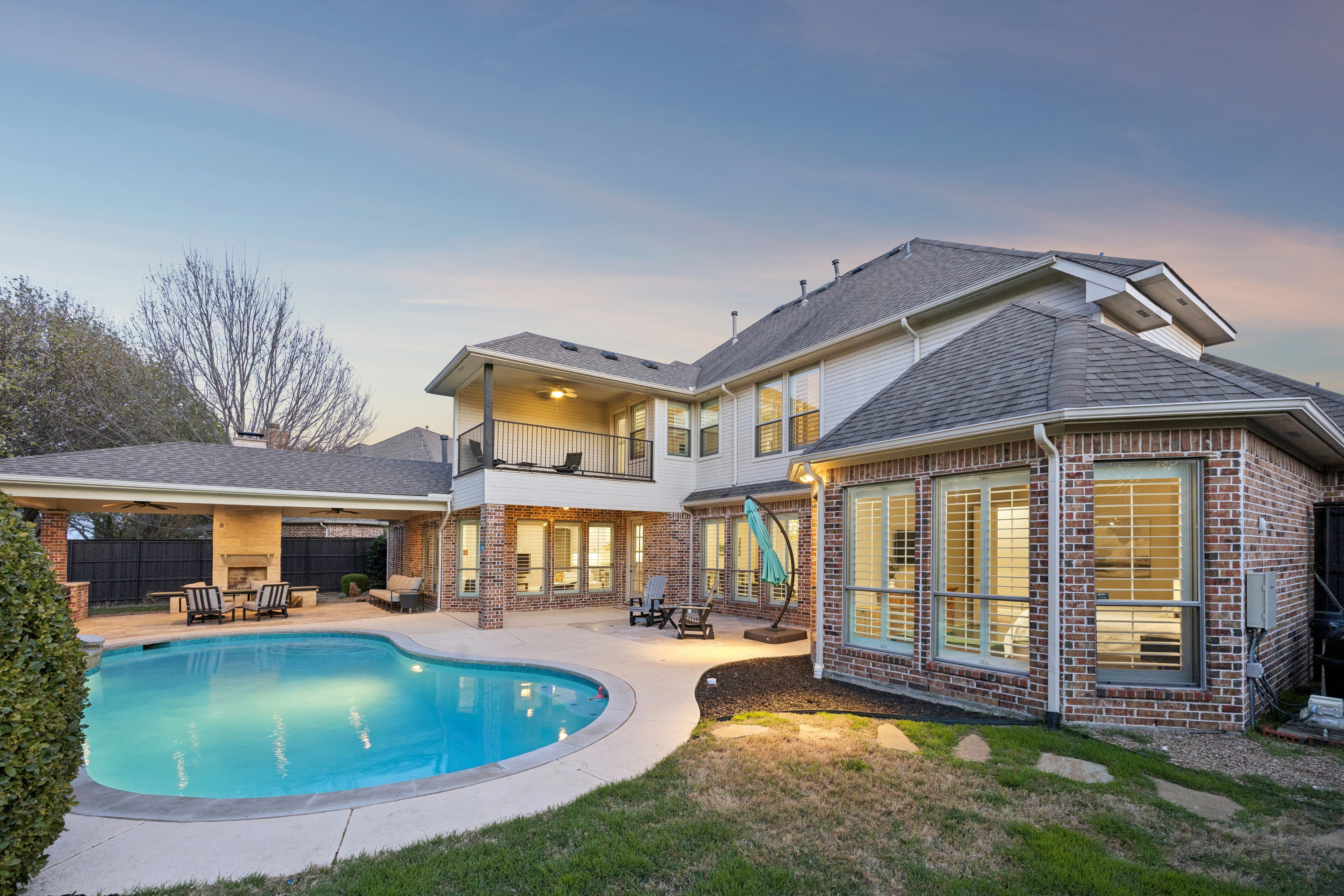 Rear exterior of a two-story brick home at dusk with a curved swimming pool, covered patio, second-story balcony, and outdoor fireplace