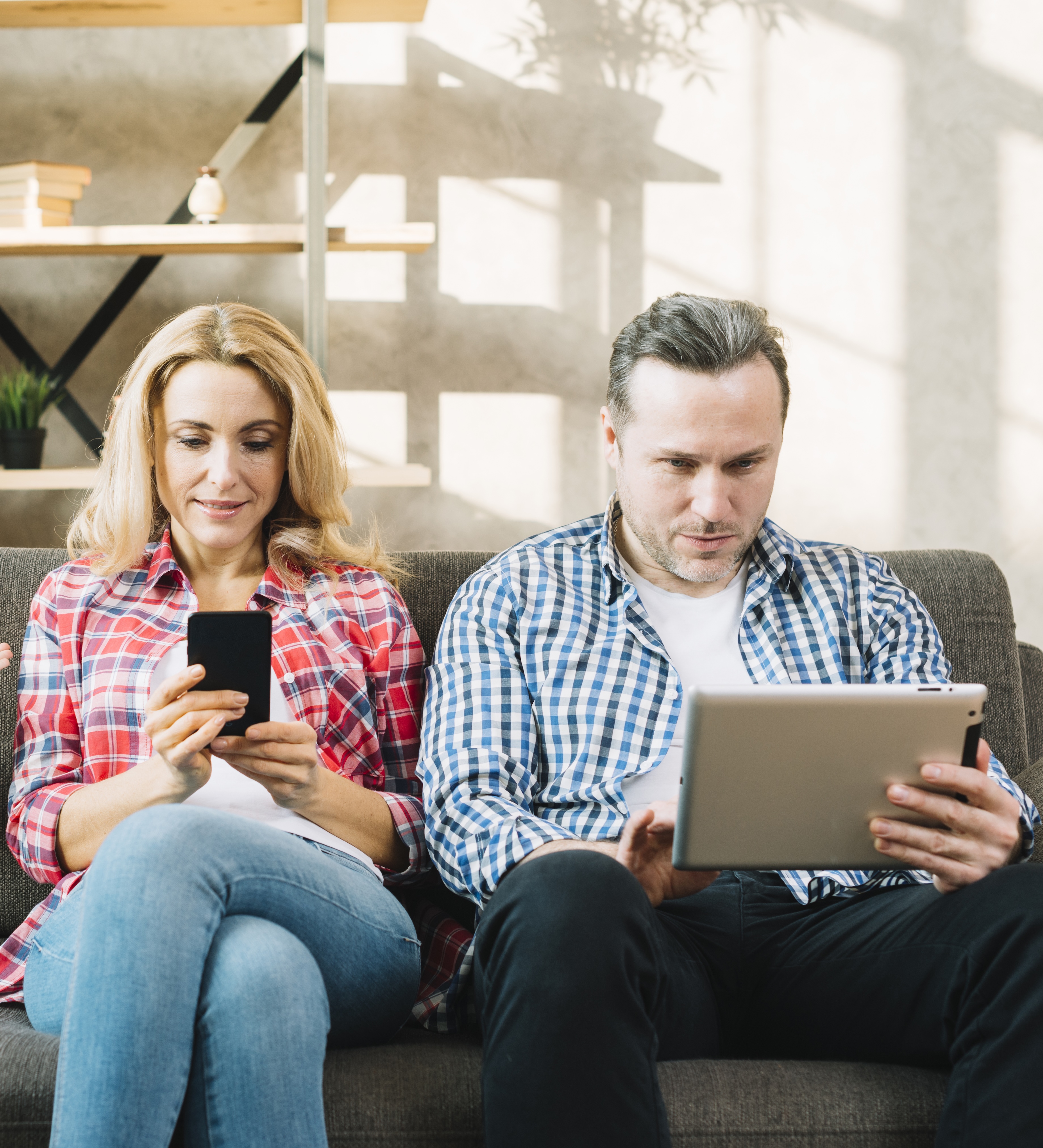 A man and a woman seated on a sofa, each looking up information on a phone and a tablet