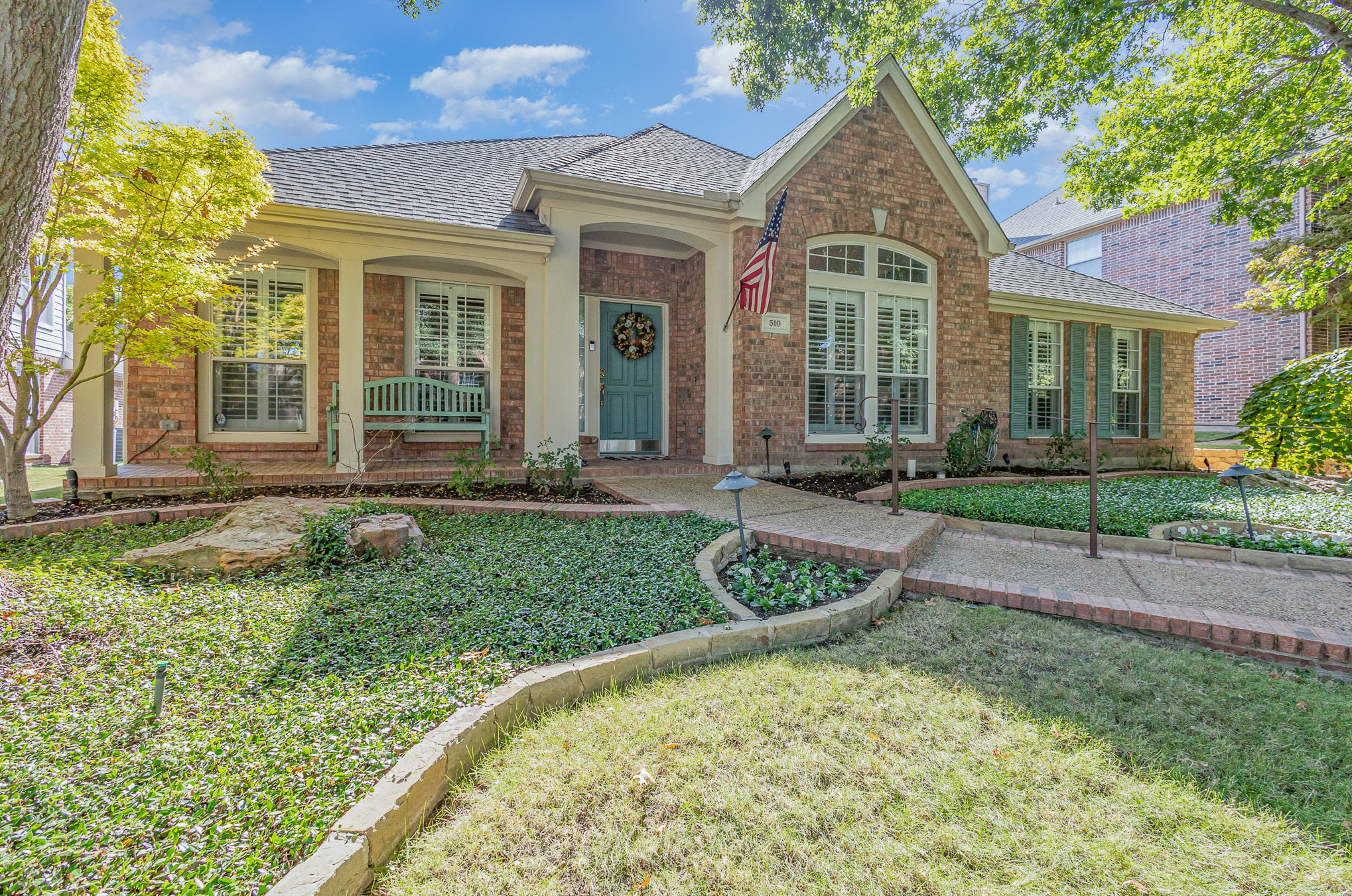 Single-story brick home with covered front porch, teal front door, and manicured landscaping in a North Texas neighborhood