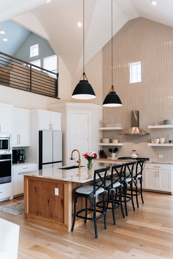 Vertical photo of a bright, vaulted-ceiling kitchen with white cabinetry, a wood-front island with marble countertop, black cross-back barstools, black dome pendant lighting, and a loft/catwalk visible above.