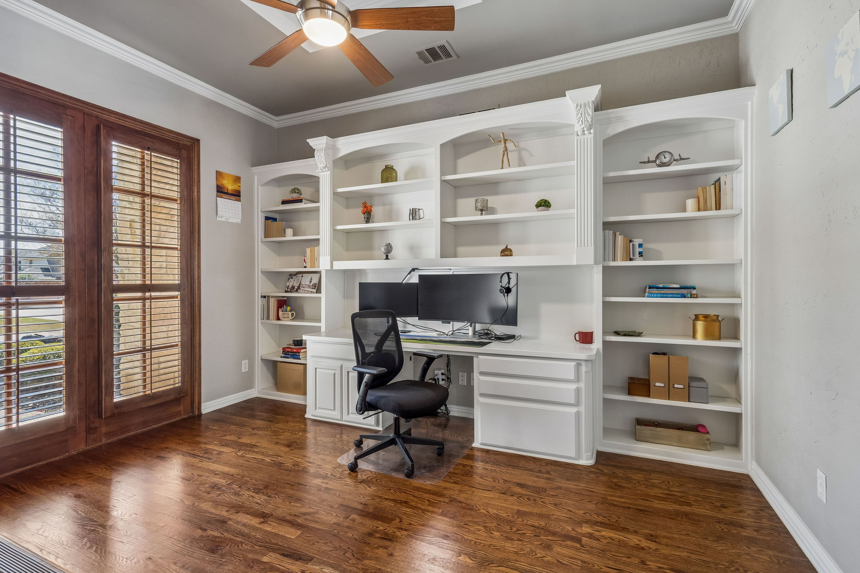 Home office with white built-in bookshelves and desk, hardwood floors, plantation shutters, and French doors
