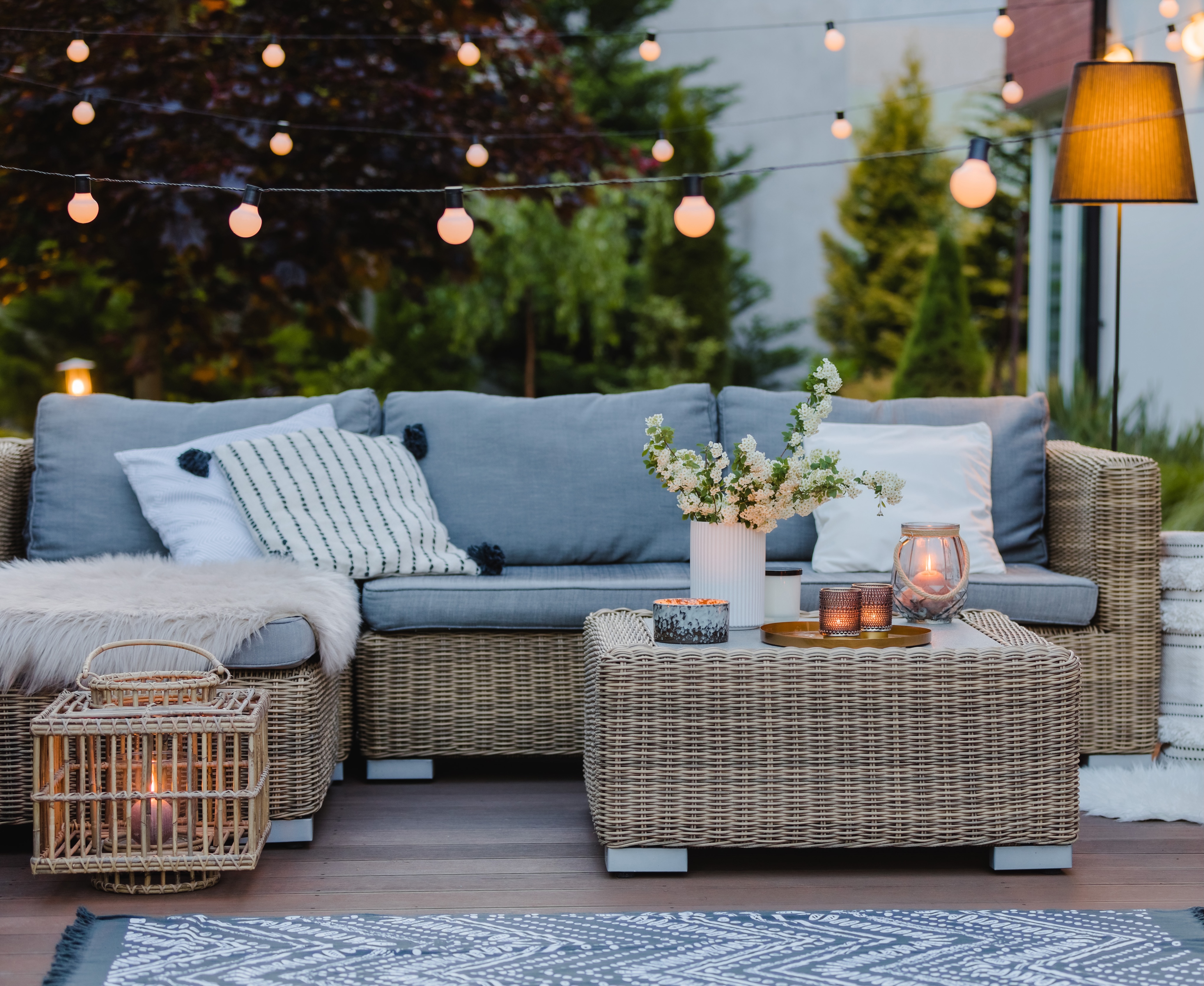Covered deck at dusk with a gray wicker sectional, cushions and throws, candle lanterns, and string lights overhead