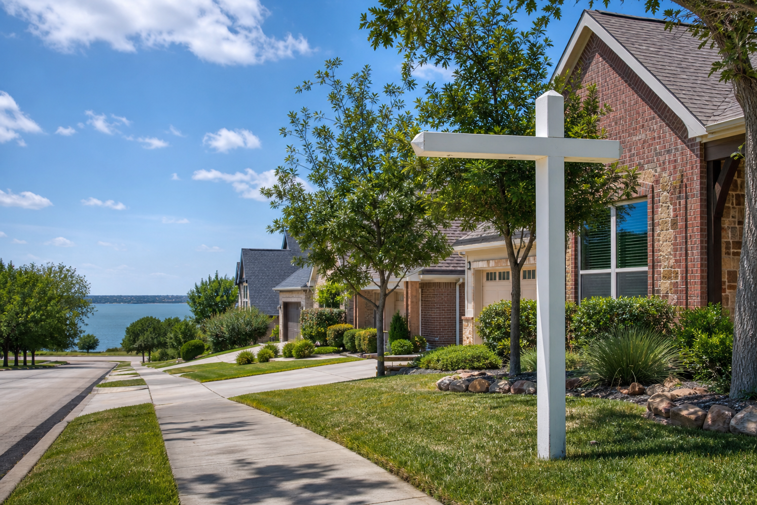 Rowlett-style residential neighborhood with brick homes, landscaped lawns, a real estate sign, and a lake view at the end of the street.