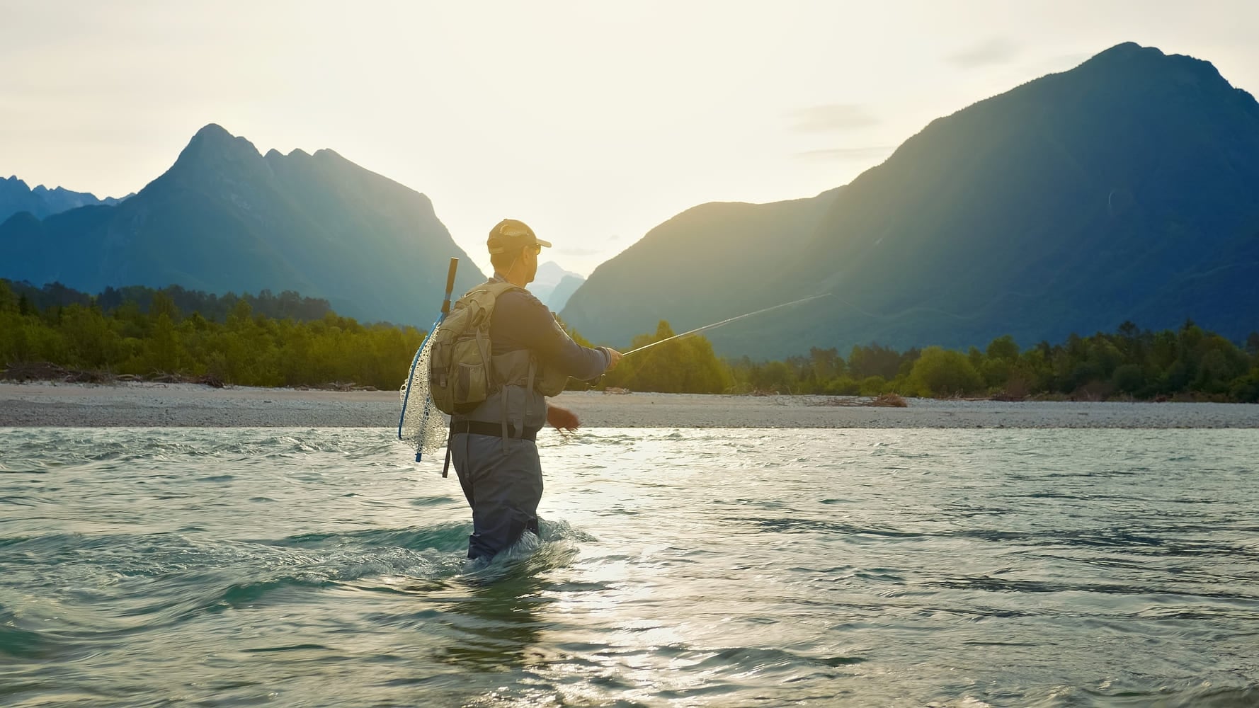 A man in outdoor gear fly-fishes in a serene river at sunset, surrounded by majestic mountains. The environmental portrait captures the tranquility and skill of freshwater fishing.
