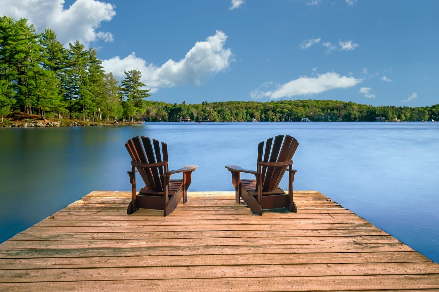Tranquil Retreat Two Adirondack chairs on a wooden dock by a serene lake. Rustic cottages nestled among lush trees dot the shoreline.