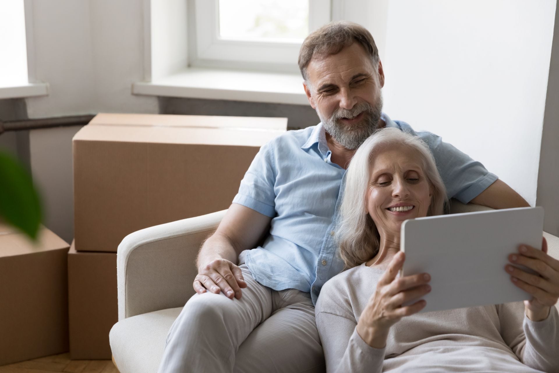Older couple relax on sofa on move day near boxes with stuff, use tablet buy goods on internet, make order via e-service app